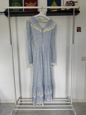 Ljusblå långklänning från Lacemade -  Ljusblå långklänning med vita broderade blommor och spetsdetaljer från Lacemade! Klänningen kan knytas i ryggen för att dra in och framhäva midjan! Här är måtten från LaceMades hemsida! Bröst: 115cm, Midja: 95cm, Höft: 120cm