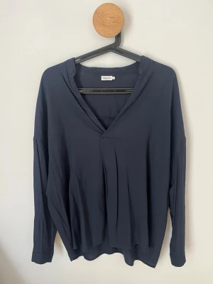 Mörkblå blus från Filippa K - Stilren mörkblå blus från Filippa K med v-ringning och avslappnad passform. Blusen har långa ärmar och ett mjukt, följsamt tyg som ger en skön känsla. Perfekt till jeans eller kjol för en enkel men snygg look.