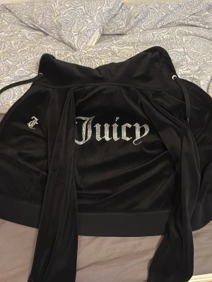 Juicy Couture svart velour hoodie - Svart hoodie från Juicy Couture i mjuk velour med glittrande strassdetaljer och stor Juicy-text på ryggen. Tröjan har huva, snörning och dragkedja med en snygg J-berlock. Perfekt för dig som gillar ikonisk Y2K-stil. Den är äkta och har använts lite, har tappat lite färg på dragkedjan men det syns knappt om man inte zoomar in. Värd 800kr men sänker priset på grund av användning. 