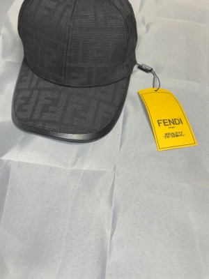 Svart keps från Fendi med FF-mönster - Snygg svart keps från Fendi med det klassiska FF-monogrammet över hela kepsen. Kepsen har böjd skärm med läderdetalj och justerbar rem bak. Tillverkad i textil med exklusiv känsla och tydlig logga på etiketten. Perfekt för dig som gillar lyxiga accessoarer.