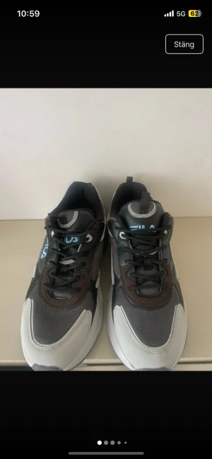Gråa Fila sneakers med bruna detaljer - Snygga sneakers från Fila i grått, svart och vitt med bruna detaljer och vit sula. Skorna har en chunky design, mesh- och syntetmaterial samt Fila-logga i blått på sidan. Perfekta för dig som gillar streetwear och sportig stil.