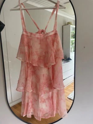 Rosa volangklänning med smala band - Söt långklänning i ljusrosa och vitt med volanger och smala axelband. Klänningen har omlottkänsla och knyt i ryggen. Luftigt material som passar perfekt till varma dagar. Mönstret är mjukt och ger en romantisk vibe.