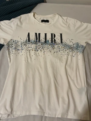 Vit AMIRI t-shirt med blått mönster - Säljer en vit t-shirt från AMIRI med coolt stänkmönster i blått och svart över bröstet och ryggen. T-shirten har korta ärmar och en klassisk passform. Perfekt för dig som vill sticka ut med en unik design.