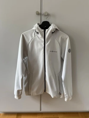 Vit vindjacka från Sail Racing - Snygg vit vindjacka från Sail Racing med huva och dragkedja framtill. Jackan har två sidofickor med dragkedja samt logga på bröstet och ärmen. Perfekt för blåsiga dagar och enkel att matcha med allt. Jackan har lite mindre fläckar på olika ställen, kan gå att få bort i tvättmaskin.