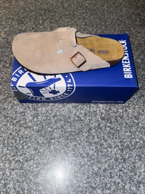 Beige mockatofflor från Birkenstock - Säljer ett par snygga beige tofflor från Birkenstock i mjuk mocka. Modellen har en bred rem med metallspänne och klassisk korksula för extra komfort. Perfekta för dig som gillar stilrena och bekväma skor till vardags.