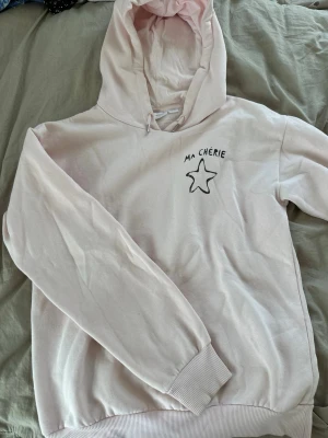 Ljusrosa hoodie med tryck från Gina tricot  - Ljusrosa hoodie från Gina tricot med huva och dragsko. På bröstet finns ett svart tryck med texten 'MA CHÉRIE' och en stjärna. Tillverkad i mjukt bomullsmaterial och har ribbade muddar vid ärmar och nederkant.
