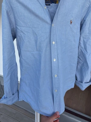 Ljusblå skjorta från Polo Ralph Lauren - Klassisk ljusblå skjorta från Polo Ralph Lauren med broderad logga på bröstet. Skjortan har knappar framtill och lång ärm. Tillverkad i mjuk bomull för en bekväm känsla och stilren look.