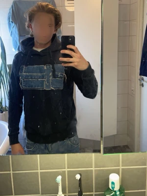 Mörkblå hoodie med jeansflagga - Säljer en mörkblå hoodie unik flaggmönster i blå denim på bröstet. Hoodien har huva med snören och ribbade muddar vid ärmslut och midja. Perfekt för dig som gillar streetwear och vill sticka ut med coola detaljer.