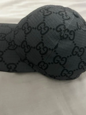 Svart keps från Gucci med GG-mönster - Snygg svart keps från Gucci med det klassiska GG-monogrammet över hela kepsen. Tillverkad i ett rutmönstrat tyg som ger en exklusiv känsla. Perfekt accessoar för att lyfta din streetwear-look.