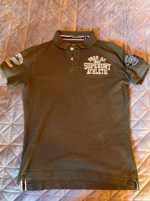 Grön Superdry pikétröja  - Snygg grön pikétröja från Superdry med broderad vit text på bröstet och tryck på båda ärmarna. Storleken är inte L enligt mig, mer mellan M och L.