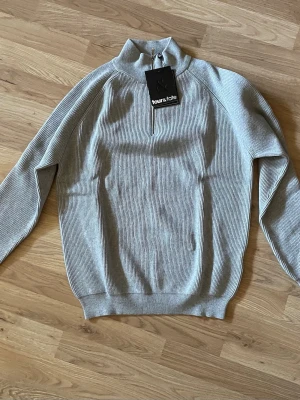 Ljusgrå stickad tröja med halvzip - Snygg ljusgrå stickad tröja från Four & Tate med halvzip och hög krage. Tröjan har ribbad struktur, långa ärmar och breda muddar vid ärmslut och nederkant. Perfekt för lager på lager och enkel att matcha med jeans eller byxor. Aldrig använd, lappen kvar. 