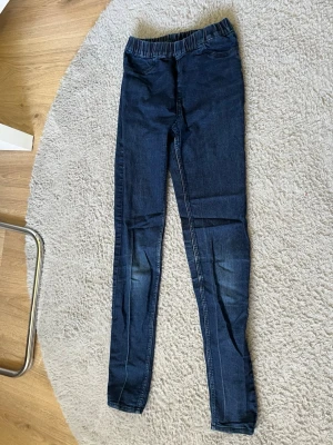 Mörkblå denimjeggings från H&M - Säljer ett par mörkblå jeggings från H&M i stretchigt denimtyg. De har resår i midjan och smal passform hela vägen ner. Perfekta för dig som gillar bekväma men ändå snygga byxor med jeanslook. Materialet är mjukt och följsamt.