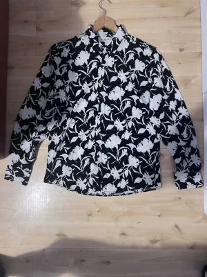 Svartvit blommig skjorta Fitmens - Snygg slim fit skjorta från Fitmens med svart botten och vita blommor. Skjortan har klassisk krage, knappar framtill och långa ärmar. Perfekt för dig som vill sticka ut med ett coolt mönster och modern passform.