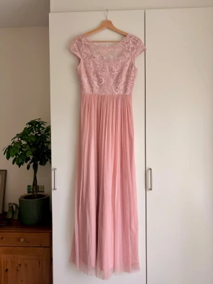 Lång rosa spetsklänning - använd 1 gång! - Romantisk långklänning i ljusrosa med spetsdetaljer upptill och transparent mesh vid halsen. Kjolen är mjukt fallande och överdelen har blommigt spetsmönster. Korta ärmar och rundad halsringning ger en elegant look. Perfekt för speciella tillfällen. Passar inte mig längre men är bra längd för mig som är ca 165 cm 🥰