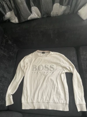 Vit stickad tröja från Hugo Boss - Vit stickad tröja från Hugo Boss med stor BOSS-logga och texten HUGO BOSS broderat på bröstet. Tröjan har rund halsringning, långa ärmar och ribbade muddar vid ärmslut och nederkant. Perfekt för en clean och stilren look.