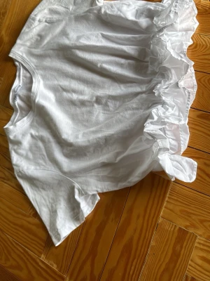 Vit t-shirt med volang från Zara - Säljer en vit t-shirt från Zara i bomull med korta ärmar och en stor volangdetalj på ena sidan. T-shirten har en klassisk rund halsringning och en clean, ljus look som sticker ut tack vare volangen.