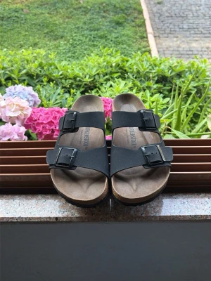 Svarta Birkenstock sandaler - Klassiska svarta Birkenstock sandaler med två breda remmar och justerbara metallspännen. Fotbädden är i kork och mocka för extra komfort och stöd. Perfekta för en avslappnad och stilren look under varma dagar.