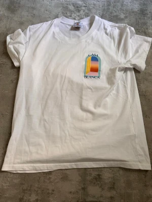 Vit Casa Blanca t-shirt med tryck - STORLEK S/M. Snygg vit t-shirt från Casa Blanca med färgglatt grafiskt tryck fram och stort tryck på ryggen. T-shirten har rund hals och korta ärmar. Perfekt för dig som gillar stilrena plagg med en twist. Tillverkad i mjuk bomull för skön känsla.