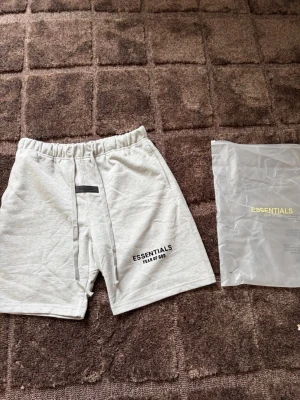 Grå Essentials Fear of God shorts - Snygga ljusgrå shorts från Essentials Fear of God med svart logga på vänster ben. De har hög midja, långa snören i midjan och är gjorda i mjuk bomull. Perfekta för en avslappnad och trendig stil.