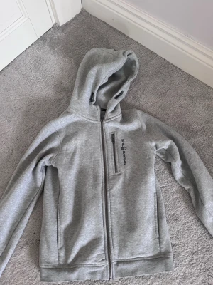 Grå hoodie från Sail Racing - Barnstorlek. Snygg grå hoodie från Sail Racing med dragkedja framtill och en bröstficka med zip. Hoodien har en klassisk huva och ribbade muddar vid ärmslut och nederkant. Tillverkad i mjukt sweatshirtmaterial, perfekt för chill dagar.