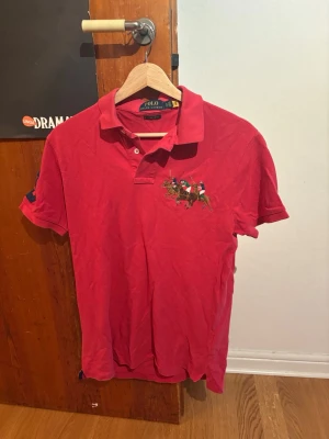Röd pikétröja från Polo Ralph Lauren - Snygg röd pikétröja från Polo Ralph Lauren med klassisk krage och knappar framtill. Broderad polospelare på bröstet och nummerdetalj på ärmen. Perfekt till jeans eller shorts för en sportig look. Nypris.: 1500kr