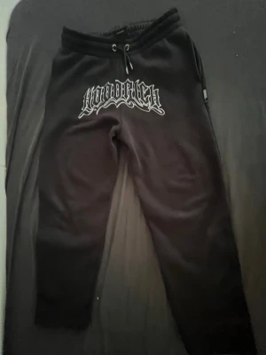 Svarta mjukisbyxor Hoodrich - Svarta mjukisbyxor från Hoodrich med vit logga i streetstil framtill. Byxorna har resår i midjan med dragsko och raka ben. Perfekta för chill dagar och har en modern, avslappnad passform. Materialet ser ut att vara mjukt och bekvämt, troligen bomullsblandning.