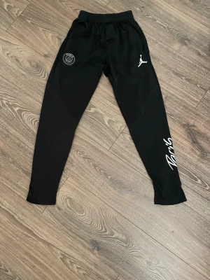 PSG träningsbyxor - Snyggt svart träningsset från PSG och Jordan med gråa detaljer och mönster på tröjan. Setet består av långärmad tröja och matchande byxor med Paris-logga och Jordan-logga. Perfekt för fotbollsträning eller chill. Materialet är troligtvis polyester för bästa komfort. Jag är 165 och den passar perfekt!