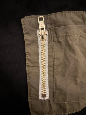Svart topp med armficka och dragkedja - Cool svart topp med en olivgrön ficka och guldfärgad dragkedja på ärmen. Fickan ger en utility-vibe och dragkedjan är en snygg detalj. Perfekt för dig som gillar streetwear och unika detaljer.