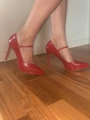 Röda lackpumps med Mary Jane-rem - Snygga röda pumps i lack med smal Mary Jane-rem och hög klack. Skorna har en spetsig tå och elegant siluett som ger en trendig look. Perfekta för dig som vill sticka ut med färg och stil. Materialet är glansigt syntetläder.