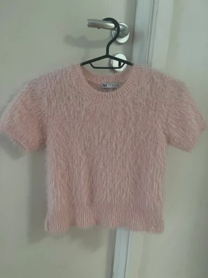 Ljusrosa fluffig stickad topp Zara - Supersöt ljusrosa stickad kortärmad topp från Zara med korta ärmar och fluffig, mjuk textur. Rund halsringning och avslappnad passform. Perfekt för dig som gillar trendiga och mysiga plagg med lite extra attityd.