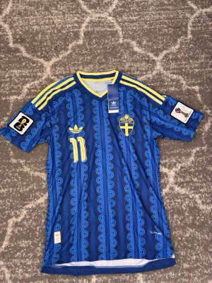 Blå Sverige fotbollströja Adidas - Säljer en blå fotbollströja från Adidas med Sveriges landslagsemblem och nummer 11 i gult. Tröjan har gula detaljer, tre ränder på axlarna och FIFA-märke på ärmen. Mönstrad design och V-ringad krage. Perfekt för dig som älskar fotboll och vill stötta Sverige.