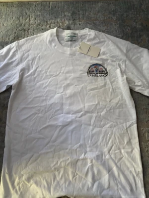 Vit t-shirt från Casablanca - Snygg vit t-shirt från Casablanca med ett färgglatt tryck av en byggnad och logga på bröstet. Klassisk rund hals och korta ärmar. Tillverkad i mjuk bomull som känns skön mot huden. Perfekt för dig som gillar stilrena plagg med en twist.
