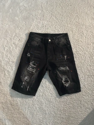 Svarta slitna jeansshorts med färgstänk - Säljer ett par svarta jeansshorts med slitna detaljer och färgstänk i blått, vitt och rött. Shortsen har klassisk femficksdesign och knappgylf. Perfekta för dig som gillar en edgy streetstil.