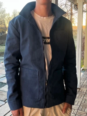 Mörkblå jacka med hög krage - Snygg mörkblå jacka med dragkedja och hög krage. Jackan har två stora fickor framtill och är tillverkad i ett slätt tyg som ger en stilren look. Passar perfekt till vardags och har en rak passform.