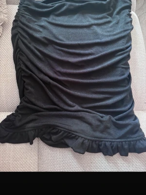 Svart volangkjol med rynkade detaljer - Trendig svart kjol med volangkant och rynkade detaljer längs sidorna. Kjolen har en kort modell och är perfekt för dig som gillar en stilren men ändå lekfull look. Tillverkad i mjukt material som ger en bekväm passform.