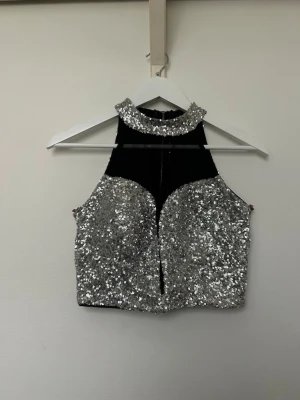 Glittrig silver croppad topp - Säljer en riktigt snygg croppad topp i svart och silver med massor av paljetter. Toppen har hög krage och är ärmlös med en cool cut-out detalj framtill. Perfekt för dig som vill sticka ut och glänsa lite extra. Inköpt på Lanzarote. Använd men i mycket fint skick. Storlek XS/S. 