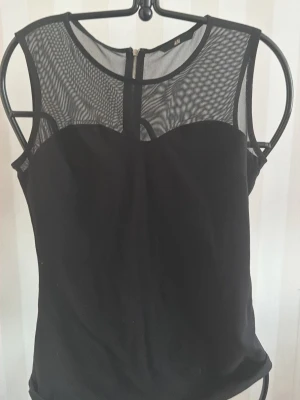 Svart mesh topp från H&M - Snygg svart topp från H&M med transparent mesh upptill och ärmlös design. Toppen har rund halsringning och dragkedja bak. Perfekt för dig som vill ha en stilren men ändå edgy look.