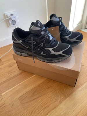 Asics Gel NYC - Snygga svarta sneakers från ASICS med grå och silverfärgade detaljer. Skorna har en chunky sula, meshpaneler för bra ventilation och klassiska ASICS-stripes på sidan. Perfekta för dig som gillar sportig och modern stil.