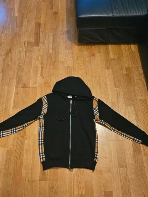Svart Burberry hoodie med rutiga detaljer - Snygg svart Burberry hoodie med dragkedja och klassiska beige-rutiga paneler längs ärmar och sidor. Huva med snören och muddar vid ärmslut och midja. Perfekt för dig som gillar streetwear och vill sticka ut lite extra.