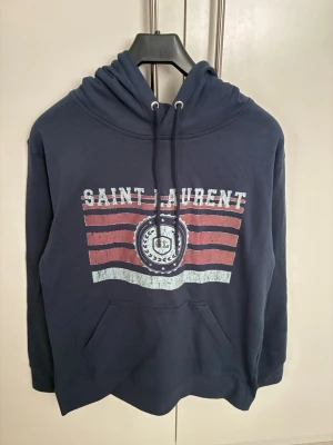 Blå Saint Laurent hoodie med tryck - Mörkblå hoodie från Saint Laurent med vit och röd vintageinspirerad logga och text på bröstet. Klassisk känguruficka framtill, justerbar huva med dragsko och ribbade muddar. Perfekt för en avslappnad och trendig look.