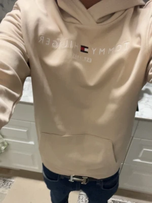 Beige hoodie från Tommy Hilfiger - Snygg beige hoodie från Tommy Hilfiger med broderad logga och flagga på bröstet. Klassisk känguruficka framtill och huva med dragsko. Perfekt för en avslappnad och trendig stil. Mjuk bomullsblandning som känns skön mot huden.