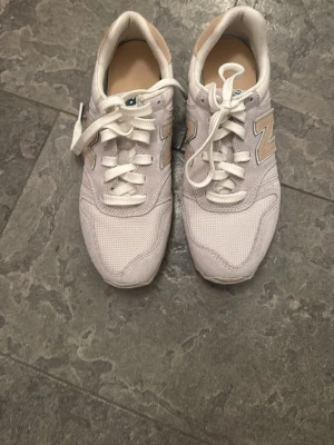 Vita New Balance sneakers med beige detaljer - Snygga vita sneakers från New Balance med beige detaljer och klassisk N-logga på sidorna. Skorna har lågt skaft, vit sula och ovandel i mesh och mocka för en sportig och fräsch look. Perfekta för dig som gillar stilrena och bekväma sneakers.