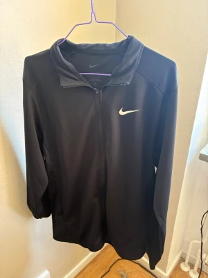 Svart träningströja från Nike - Snygg svart träningströja från Nike med dragkedja framtill och klassisk vit Nike-logga på bröstet. Tillverkad i ett lätt och funktionellt material som passar perfekt till träning eller uppvärmning. Jackan har hög krage och långa ärmar.