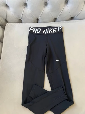 Svarta Nike Pro leggings - Säljer ett par svarta Nike Pro leggings med bred resår i midjan där det står 'PRO NIKE' i vitt. De har en tight passform och är tillverkade i ett stretchigt funktionsmaterial som passar perfekt till träning. Klassisk Nike Swoosh-logga på benet.