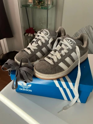 Grå Adidas campus 00s, dam, storlek 38 - Snygga grå sneakers från Adidas med klassiska vita ränder på sidorna och vit sula. Skorna är i mocka och har Adidas logga på plösen. Levereras med både vita och nya gråa skosnören. 