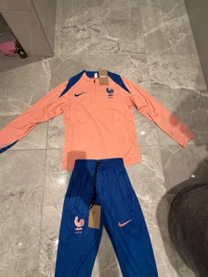 Frankrike Nike träningsset blå/korall - Snyggt träningsset från Nike med Frankrikes landslagslogga. Tröjan är långärmad i korall med blå detaljer och dragkedja vid halsen. Byxorna är blå med korallfärgade detaljer och resår i midjan. Perfekt för fotbollsträning eller chill.