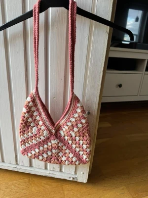 Rosa och beige virkad axelväska - Unik handvirkad axelväska i rosa, korall och vitt. Väskan har en trekantig form med axelrem och ett snyggt rutmönster. Perfekt för dig som gillar boho-stil och vill ha något personligt. Materialet är akrylgarn