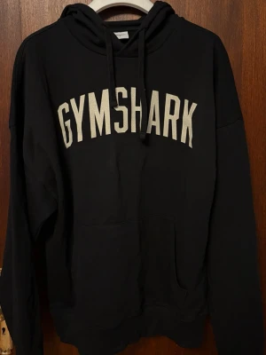 Svart Gymshark hoodie  - Snygg svart hoodie från Gymshark med stort vitt tryck över bröstet. Klassisk känguruficka framtill och justerbar huva med snören. Tillverkad i mjukt bomullsmaterial som är perfekt för chill eller gymmet. Oversized fit för en avslappnad stil.