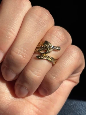 Liten Guldfärgad Ring med Fjäderdesign - Unik guldfärgad ring formad som en fjäder som slingrar sig runt fingret. I fint skick. Ingen stämpel.