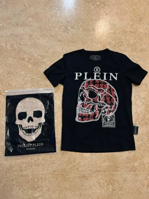 Svart Philipp Plein t-shirt med dödskalle - Snygg svart t-shirt från Philipp Plein med stor dödskalle framtill i vitt och rött samt tydlig logga. T-shirten har rund hals och normal passform. Helt ny, aldrig använd. Storlek XS. Jag  är 169cm väger 67kg och den sitter lite tajt på mig. 